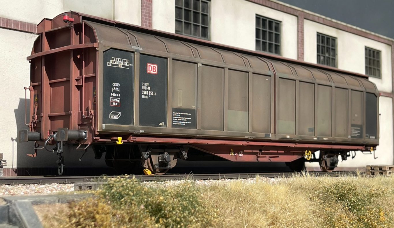 Rostiger Güterwagen an einer Bahnhofsanlage mit großen Fenstern und leerem Wageninneren.