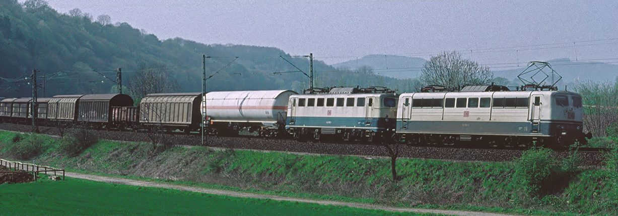 Zug fährt entlang einer ländlichen Strecke mit grünen Wiesen und Hügeln im Hintergrund.