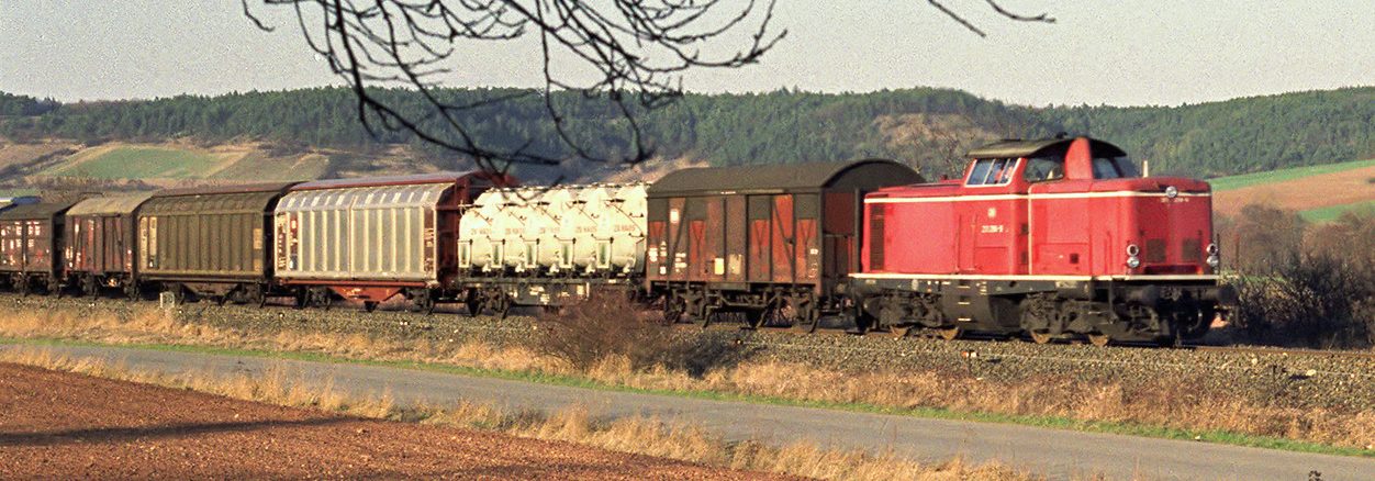 Rote Lokomotive mit Güterwagen fährt durch eine ländliche Landschaft.