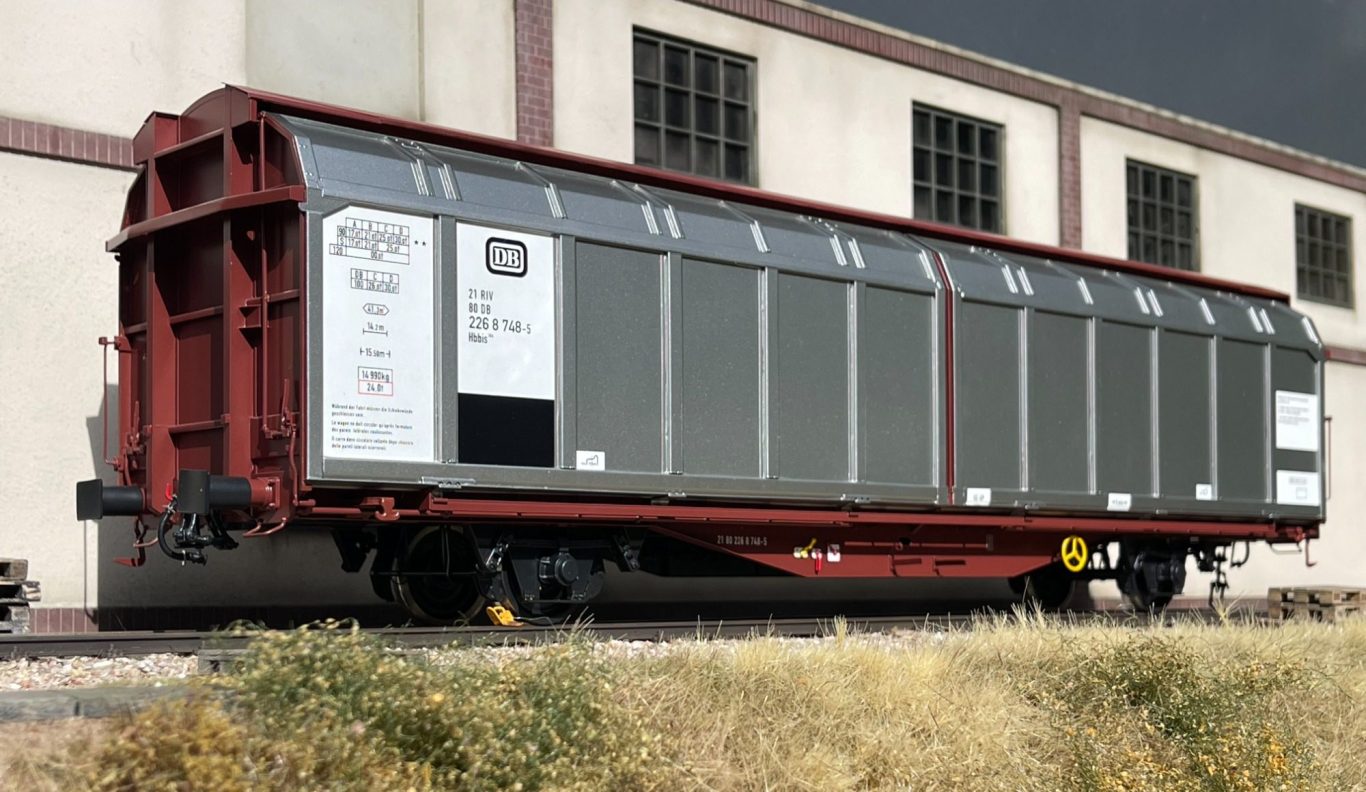 Grauer Güterwagen mit rotem Rahmen, abgestellt vor einer Fabrikwand.